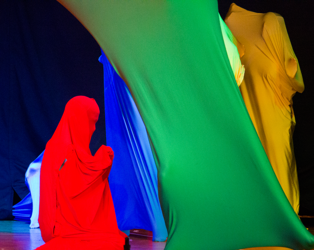 Es sind 4 Schüler*innen verborgen in elastischen Tanzsäcken zu sehen. Die Tanzsäcke haben die Farben rot, blau, grün und gelb. Schüler*in 1 in rotem Tanzsack kniet mit gefalteten Händen links im Bild, Blickrichtung nach rechts. Im Vordergrund springt ein*e Schüler*in mit grünem Tanzsack getreckt diagonal in die Höhe. Die Bewegung verläuft dynamisch von rechts unten nach links oben im Bild. Dabei verdeckt er zum Teil die beiden Schüler*innen im gelben und blauen Tanzsack. Der blaue Tanzsack ist zwischen dem roten und grünen Tanzsack im Hintergrund teilweise sichtbar. Er steht gestreckt auf Zehenspitzen. Der gelbe Tanzsack ist rechts von dem grünen im Hintergrund in aufrechter Haltung und mit gehobenen Händen sichtbar. Die Umrisse von Gesicht und Händen sind erkennbar.  Den Hintergrund bildet ein schwarzer Vorhang, vor dem die Ausschnitte zweier weiterer blauer Tanzsäcke, jedoch zum Großteil von dem Vierergespann verborgen, erkennbar sind.